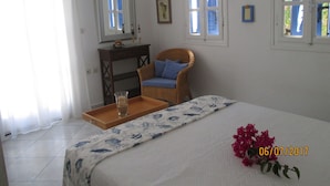 Une chambre à coucher avec un lit, un fauteuil en osier, une petite table avec un plateau et une bougie, et une fenêtre avec des volets bleus.