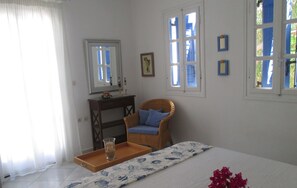 A bedroom with a bed, a wicker chair, a small table with a tray and a candle, and a window with blue shutters.