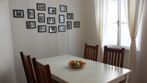 Ein Essbereich mit einem weißen Tisch, Holzstühlen und einer Obstschale. An der Wand hängen Bilder und es gibt ein Fenster mit Vorhängen.