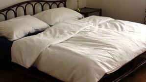Ein Bett mit hölzernem Kopfteil, weißer Bettdecke und zwei Kissen. Ein kleiner Nachttisch mit Lampe und einem dekorativen Gegenstand steht daneben.