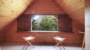 Una stanza mansardata con pareti in legno e un soffitto spiovente, con un'ampia finestra che si affaccia su una foresta. Sono presenti due sedie pieghevoli in legno.
