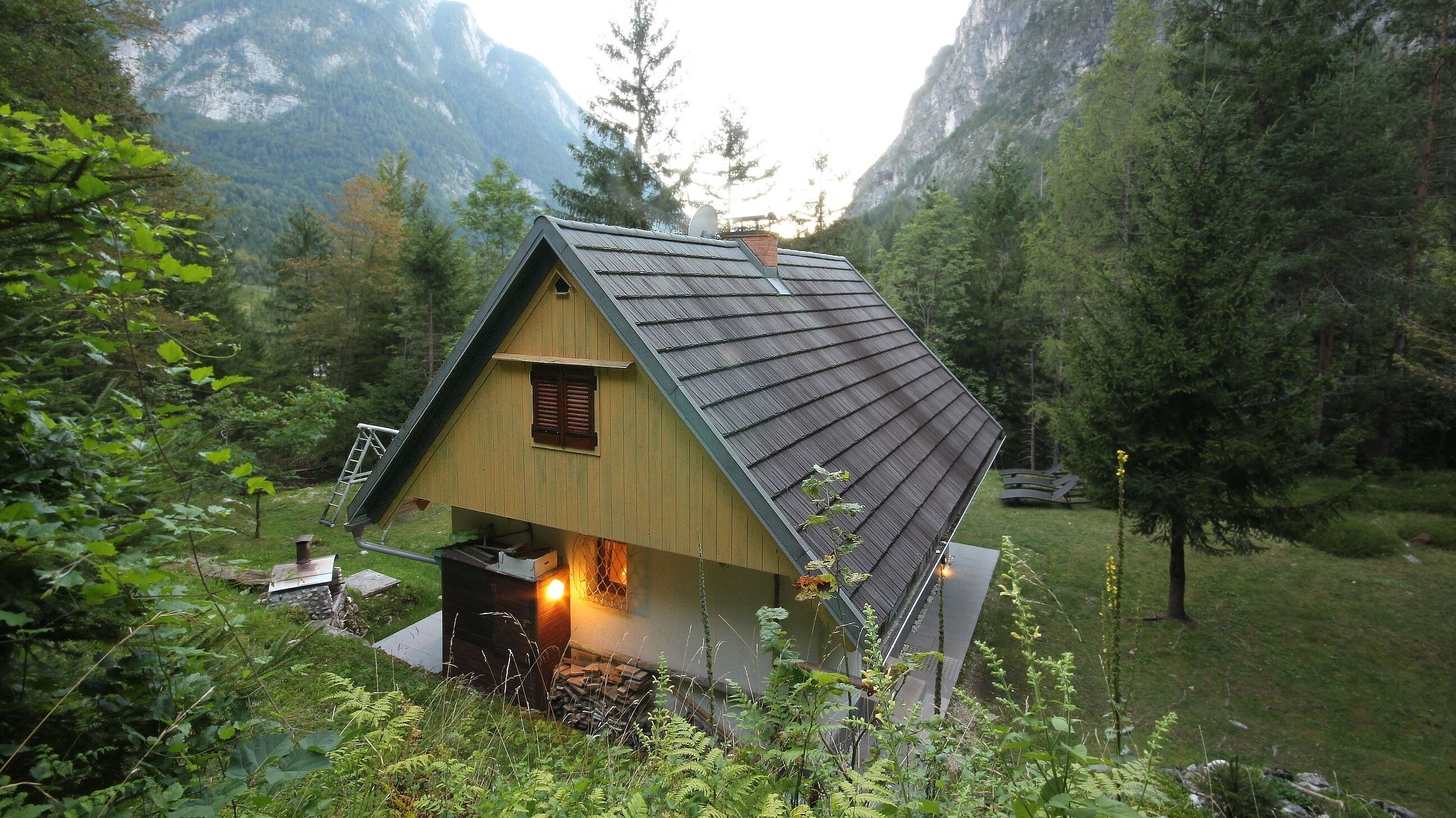 Une petite cabane avec un toit en pente, une cheminée et un intérieur éclairé, entourée d’une forêt dense et de montagnes.