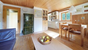 Un accogliente interno di una baita con soffitto in legno, zona pranzo con tavolo e sedie, angolo cottura con mobili e forno a microonde, e soggiorno con divano e un cesto di frutta sul tavolo.
