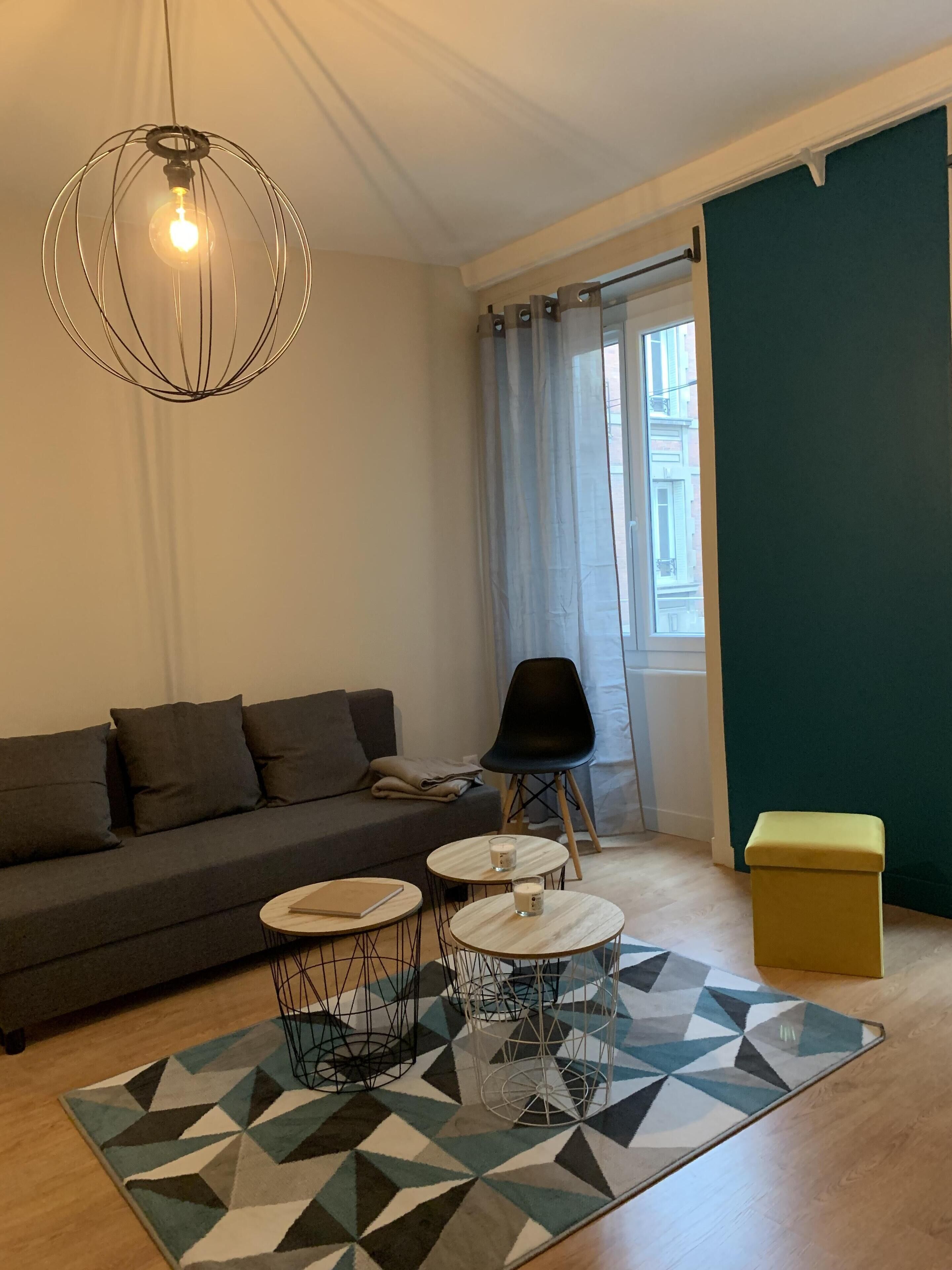 Een moderne woonkamer met een grijze bank, twee ronde tafels met krukken, een vloerkleed met een geometrisch patroon, een zwarte stoel en een gele poef.