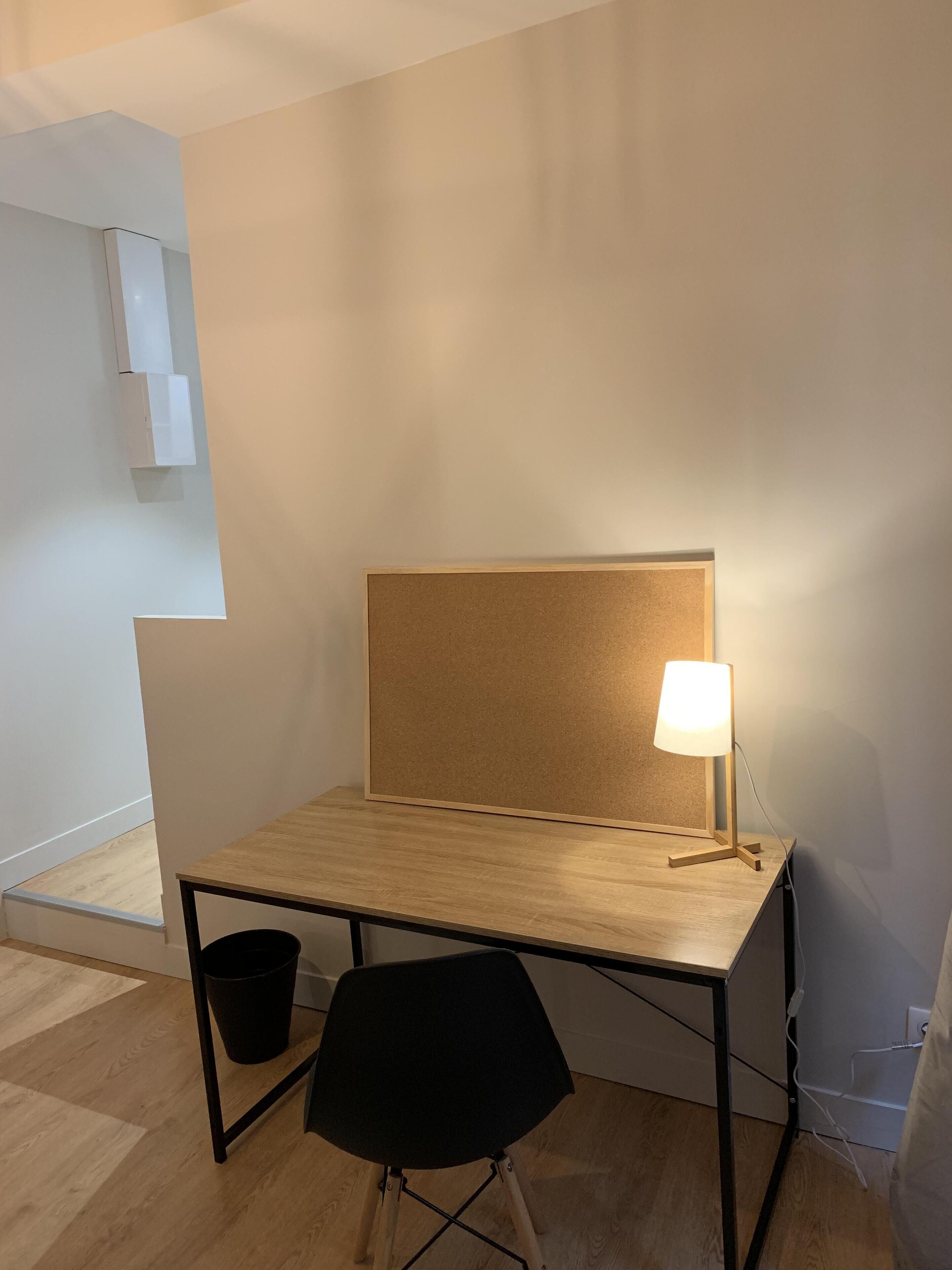 Um espaço de trabalho pequeno e bem organizado, com um quadro de cortiça, uma luminária de mesa e uma cadeira preta.