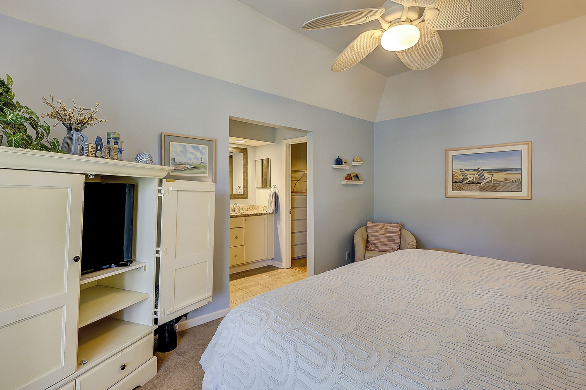 A bedroom with a bed, a wardrobe, a ceiling fan, a bathroom, and a beach-themed picture on the wall.