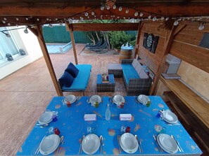 Un espace de restauration en plein air avec une nappe bleue, des meubles en bois et un coin salon couvert agrémenté de guirlandes lumineuses.