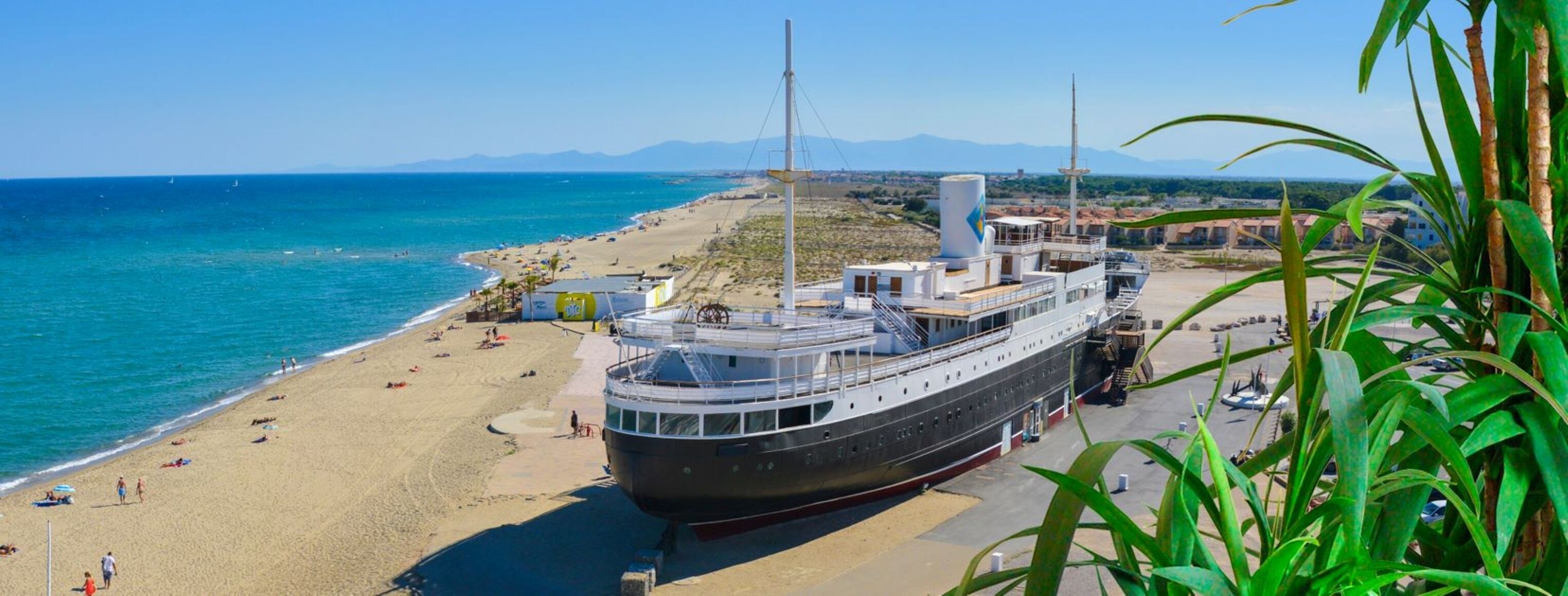 A large cruise ship docked at a beach with a clear view of the ocean and mountains in the background.