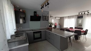 A modern kitchen with a central island, a dining area with a red tablecloth, and a living space with a TV and decorative items. - 4-STAR VILLA TORREILLES SEASIDE RESORT (TORREILLES)
