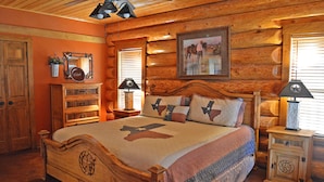Een slaapkamer in blokhutstijl met een houten bed, een nachtkastje, een dressoir en een raam met jaloezieën.