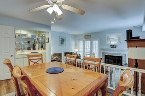 A dining room with a wooden table and chairs, a ceiling fan, a fireplace, and a kitchen area with shelves and a mirror.