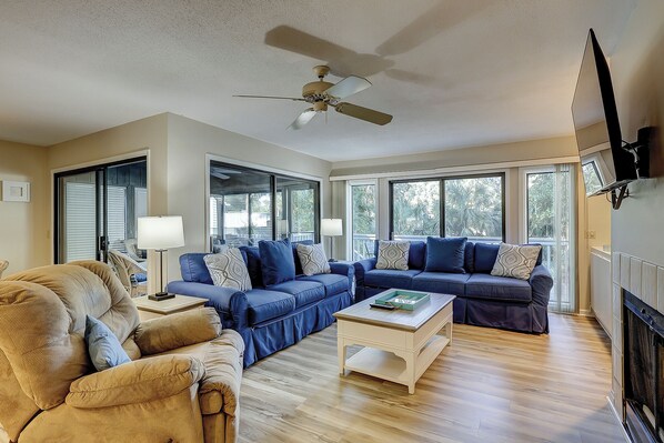 A spacious living room with a beige sofa, a blue sectional, and a white coffee table. There is a ceiling fan, large windows, and a fireplace.