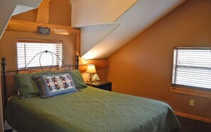 A bedroom with a sloped ceiling, a bed with a green quilt, a decorative pillow, a nightstand with a lamp, and a window with blinds.