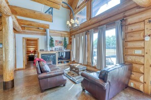 L’intérieur d’une cabane en rondins, avec une cheminée, des canapés en cuir, un téléviseur et une grande fenêtre agrémentée de rideaux.