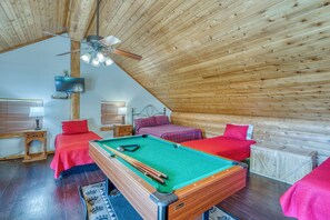 A cozy attic room with a pool table, two beds with red bedding, wooden walls, and a ceiling fan. - Private Pool, Hot Tub & Games Near Lake LBJ | Pet Friendly, Sleeps 14 (Burnet)