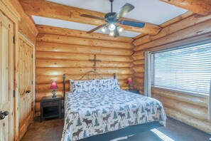 Un dormitorio tipo cabaña de troncos con una cama, dos lámparas de noche, un ventilador de techo y una ventana con persianas.