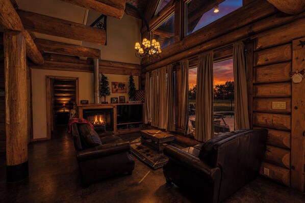 A log cabin interior with a fireplace, leather sofas, and a large window overlooking a sunset.
