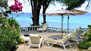 A sunlit patio with lounge chairs and a large umbrella, overlooking a beach with a clear view of the ocean. - Secret Cove 4 (Derricks)