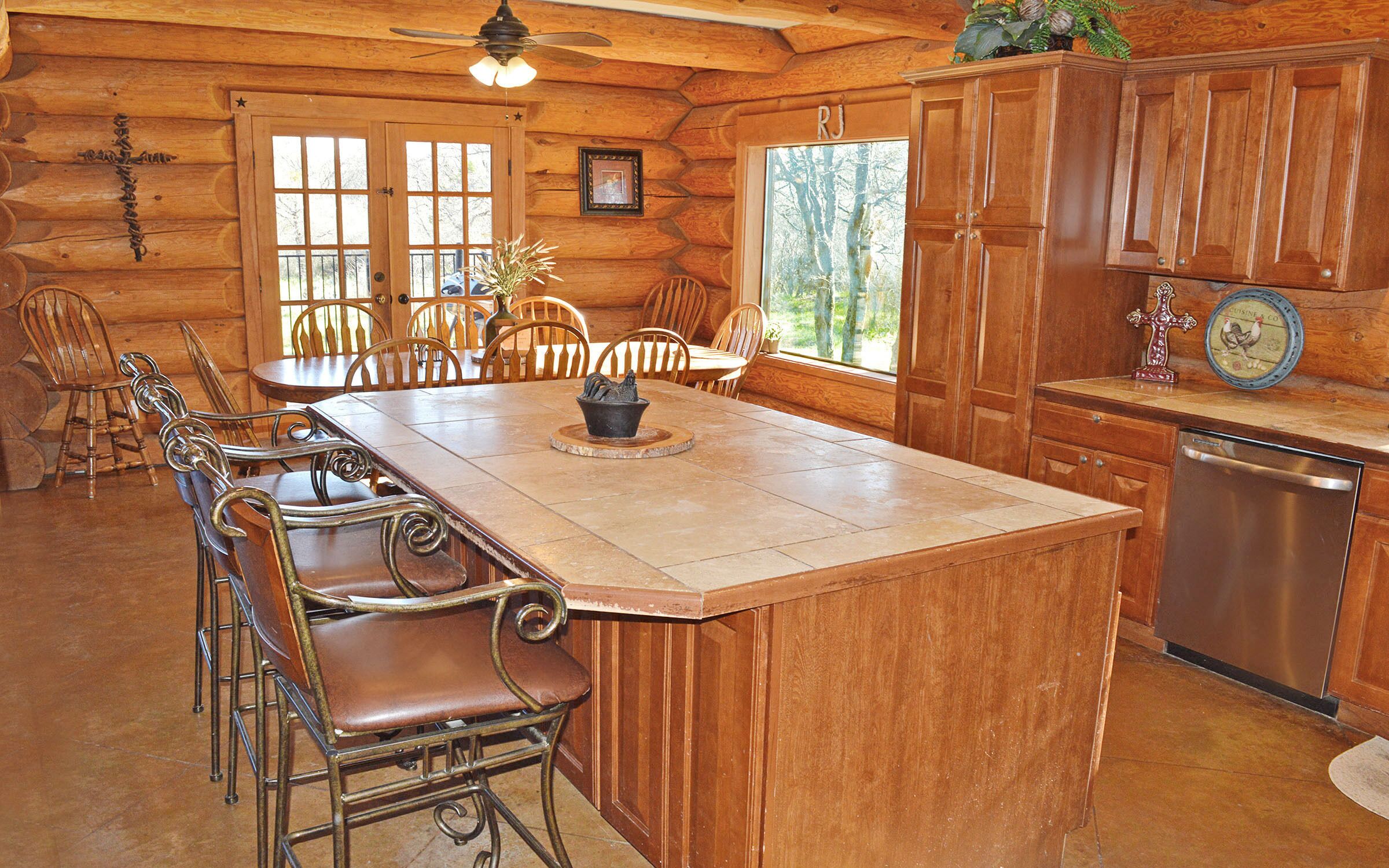 A wooden cabin interior with a large dining table, chairs, a kitchen area, and a view of the outdoors.