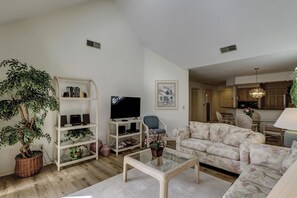 A living room with a sofa, a glass coffee table, a TV, and a bookshelf. - Sunset Rentals | Colonnade Club 168 (Hilton Head Island)