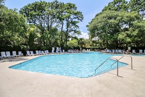 Uma piscina retangular com deck de concreto, rodeada por espreguiçadeiras brancas e árvores.