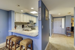 Una cocina con gabinetes blancos, encimera de granito y microondas. Hay tres taburetes de mimbre y un cartel que dice 'I love you to Hilton Head'.