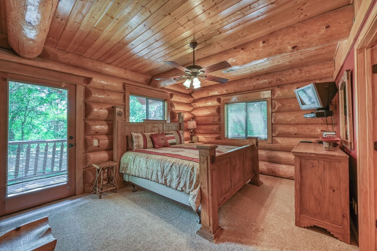 A log cabin bedroom with a bed, a nightstand, a dresser, a ceiling fan, and large windows.