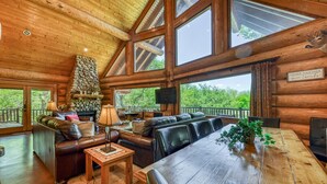 A log cabin interior with a fireplace, leather seating, a dining table, and large windows offering a view of the outdoors.