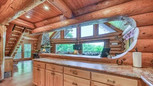 A log cabin interior with a wooden staircase, a stone fireplace, and a large window overlooking greenery.