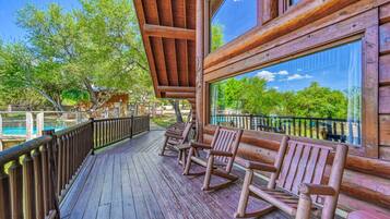 A wooden deck with rocking chairs and a glass railing overlooking a pool and trees.