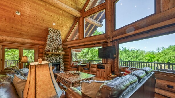 A log cabin interior with a stone fireplace, leather seating, and large windows overlooking greenery.