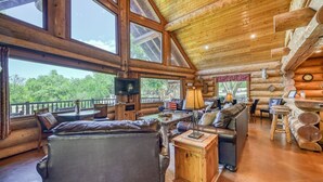 A log cabin interior with large windows, a fireplace, leather furniture, and a wooden dining table.