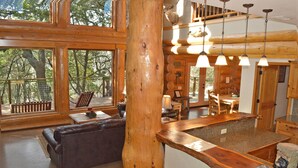 Interior de una cabaña de troncos con un ventanal grande, una mesa de comedor de madera y un sofá de cuero.
