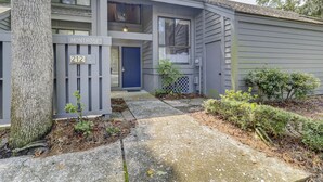 Un immeuble résidentiel de deux étages, à l’extérieur gris, avec une porte bleue et un petit jardin devant, agrémenté de plantes et d’un chemin en béton.