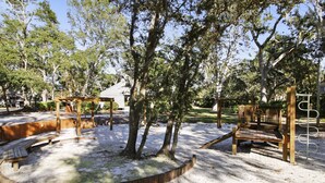 Une aire de jeux avec un bac à sable circulaire, des structures de jeu en bois et des arbres.