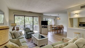 Un vaste espace de vie comprenant un canapé, une table basse, une table à manger avec des chaises, un téléviseur et un balcon doté de grandes fenêtres.
