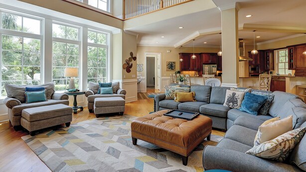 A spacious living room with a high ceiling, large windows, and a view of the outdoors. There is a kitchen area with dark wooden cabinets and a dining area with a table and chairs. - Sunset Rentals | Egret 5 (Hilton Head Island)