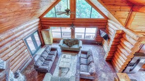 L’intérieur d’une cabane en rondins, avec un plafond haut, un ventilateur de plafond, une cheminée et une grande fenêtre.
