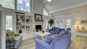 A spacious living room with a large window, a fireplace, and a bookshelf filled with books and decorative items. - Sunset Rentals | Baynard Park 6 | Dog Friendly (Hilton Head Island)