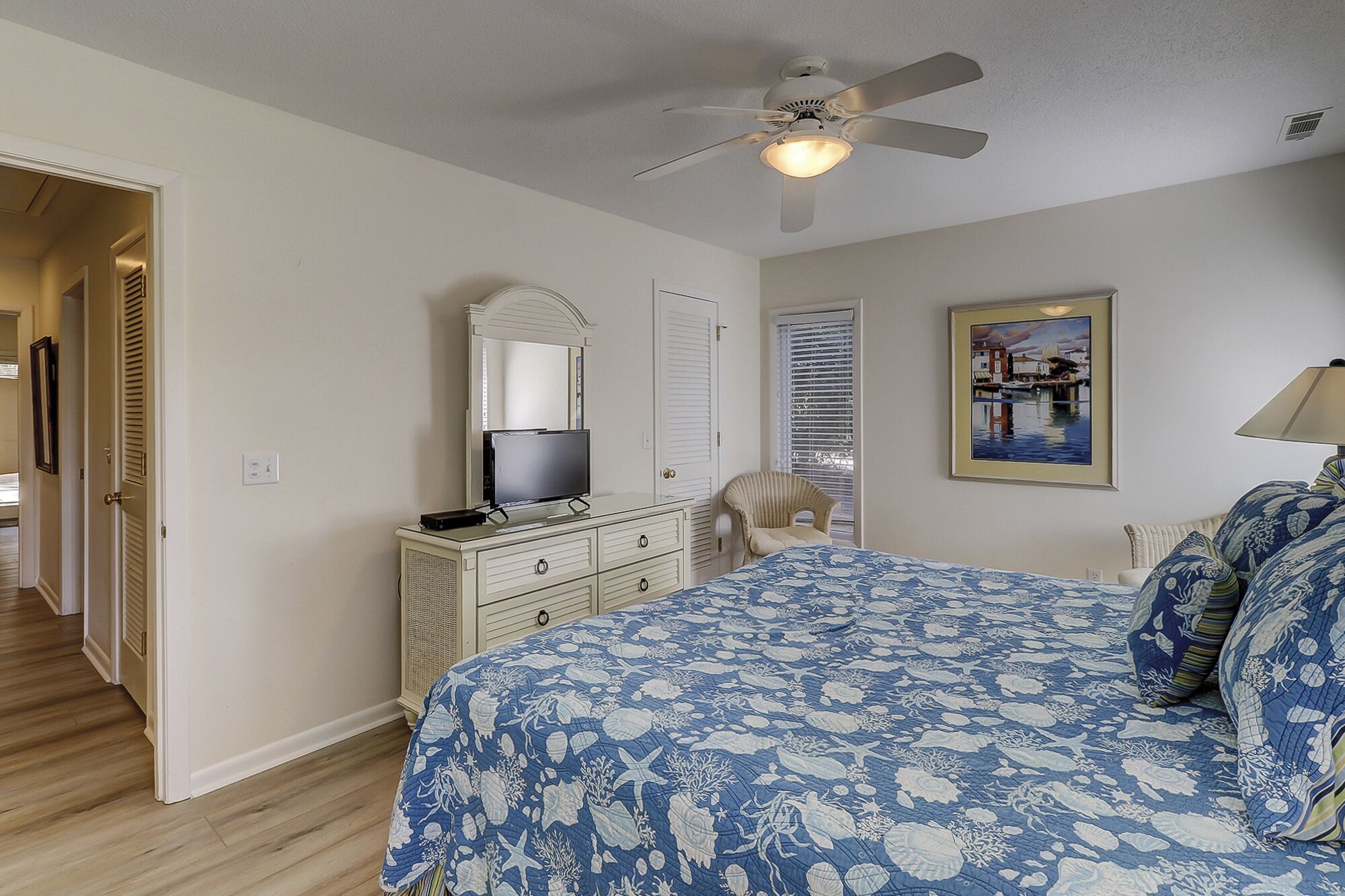 A bedroom with a bed, a dresser, a television, a ceiling fan, a window with blinds, and a framed picture on the wall.
