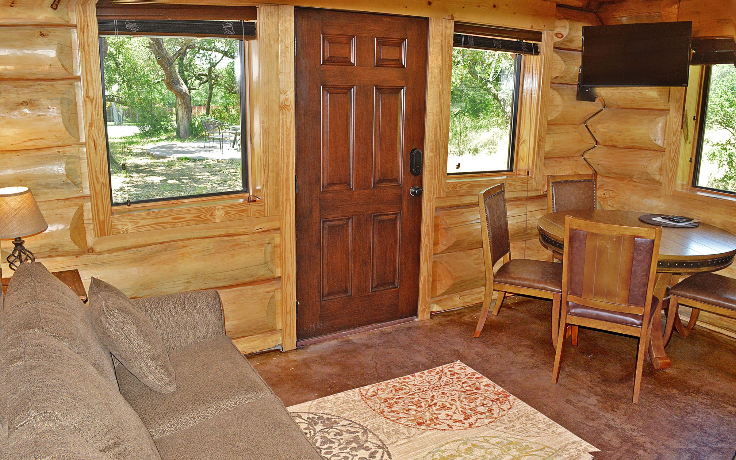 A log cabin interior with a wooden door, a dining table with chairs, a sofa, and a rug.