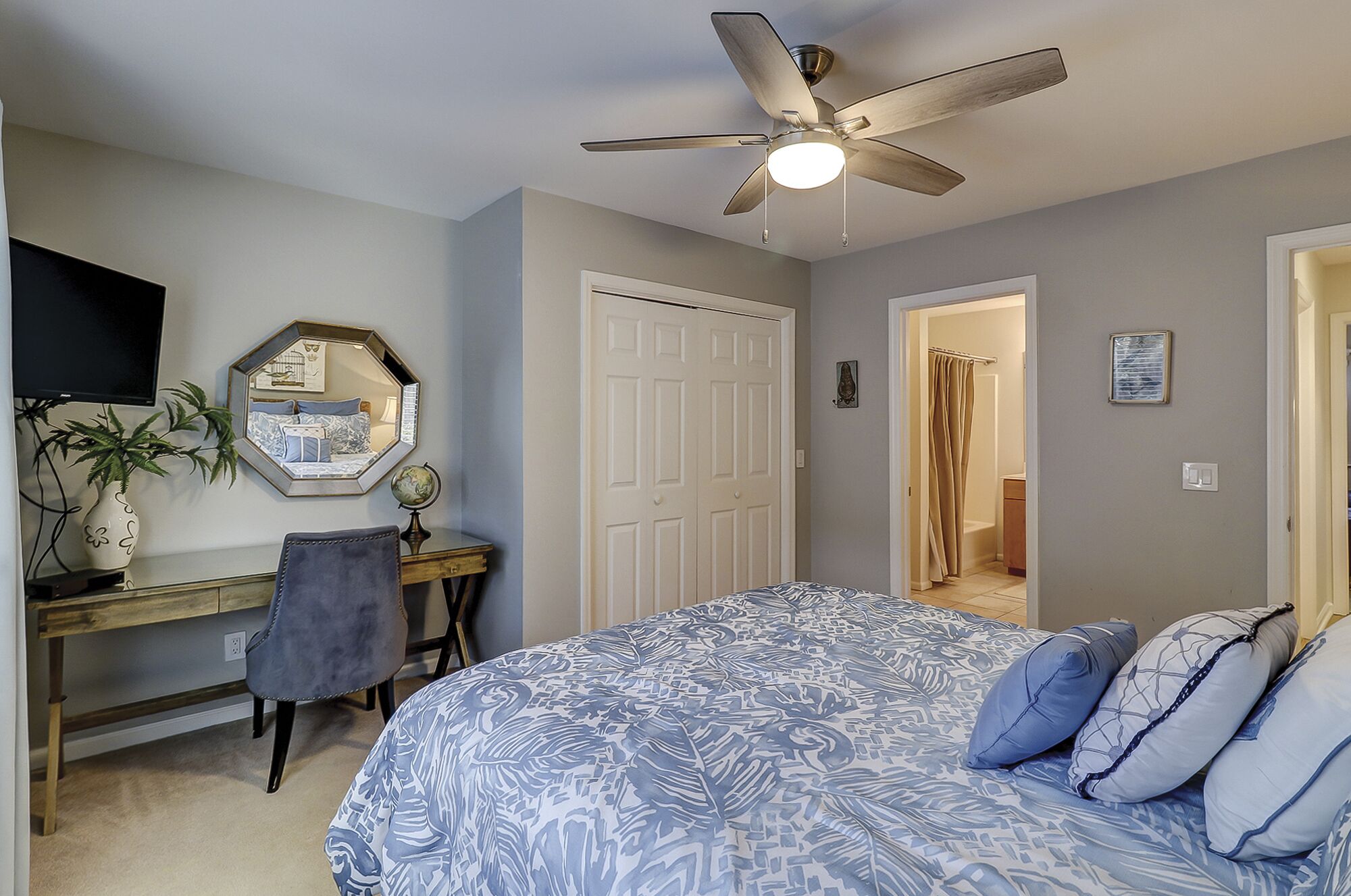 A bedroom with a bed, a desk, a chair, a ceiling fan, a mirror, and a door leading to a bathroom.