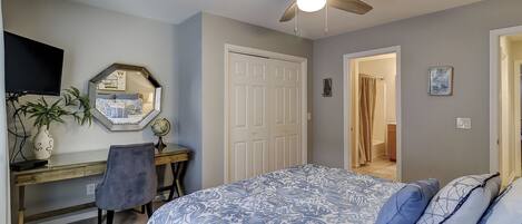 A bedroom with a bed, a desk, a chair, a ceiling fan, a mirror, and a door leading to a bathroom.
