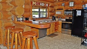 Une cuisine rustique avec un bar en bois, des tabourets, un réfrigérateur et une enseigne sur laquelle on peut lire « MATADOR ».