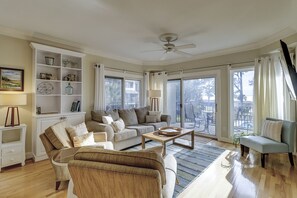 A spacious living room with a sofa set, a coffee table, and a wicker chair. There is a bookshelf with decorative items and a ceiling fan.