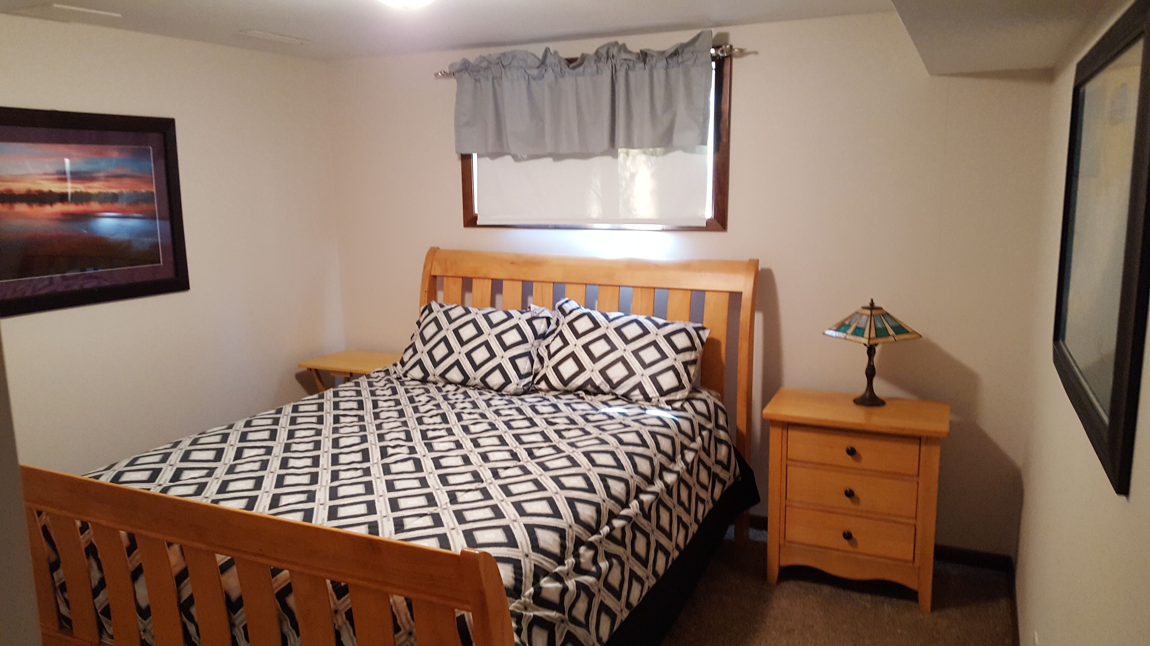 A bedroom with a wooden bed, patterned bedding, a nightstand with a lamp, a window with a curtain, and a framed picture on the wall.