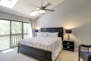 Une chambre avec un grand lit, un ventilateur de plafond, une table de chevet avec une lampe, une chaise et une vue sur la verdure par la fenêtre.