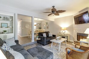 Un salon avec un canapé de couleur gris foncé, une table basse et un fauteuil en osier. On y trouve un ventilateur de plafond, un téléviseur à écran plat fixé au mur, et un coin repas visible au fond de la pièce.