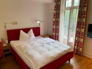 A hotel room with a large bed, a nightstand, a bedside table, a window with curtains, and a wall-mounted TV.