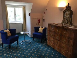 A room with two blue armchairs, a small round table, a wooden dresser with a clock, and a window with curtains.